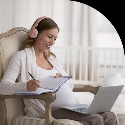 Pregnant woman with headphones writing in notebook using a laptop